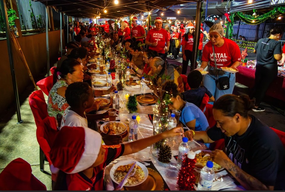Voluntários de camiseta vermelha e gorro de Papai Noel servem a ceia do Natal Invisível em uma longa mesa decorada, à noite.