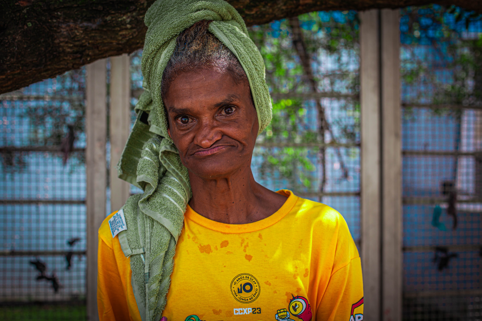 Retrato de uma mulher de camiseta amarela e toalha verde na cabeça, sob uma árvore.
