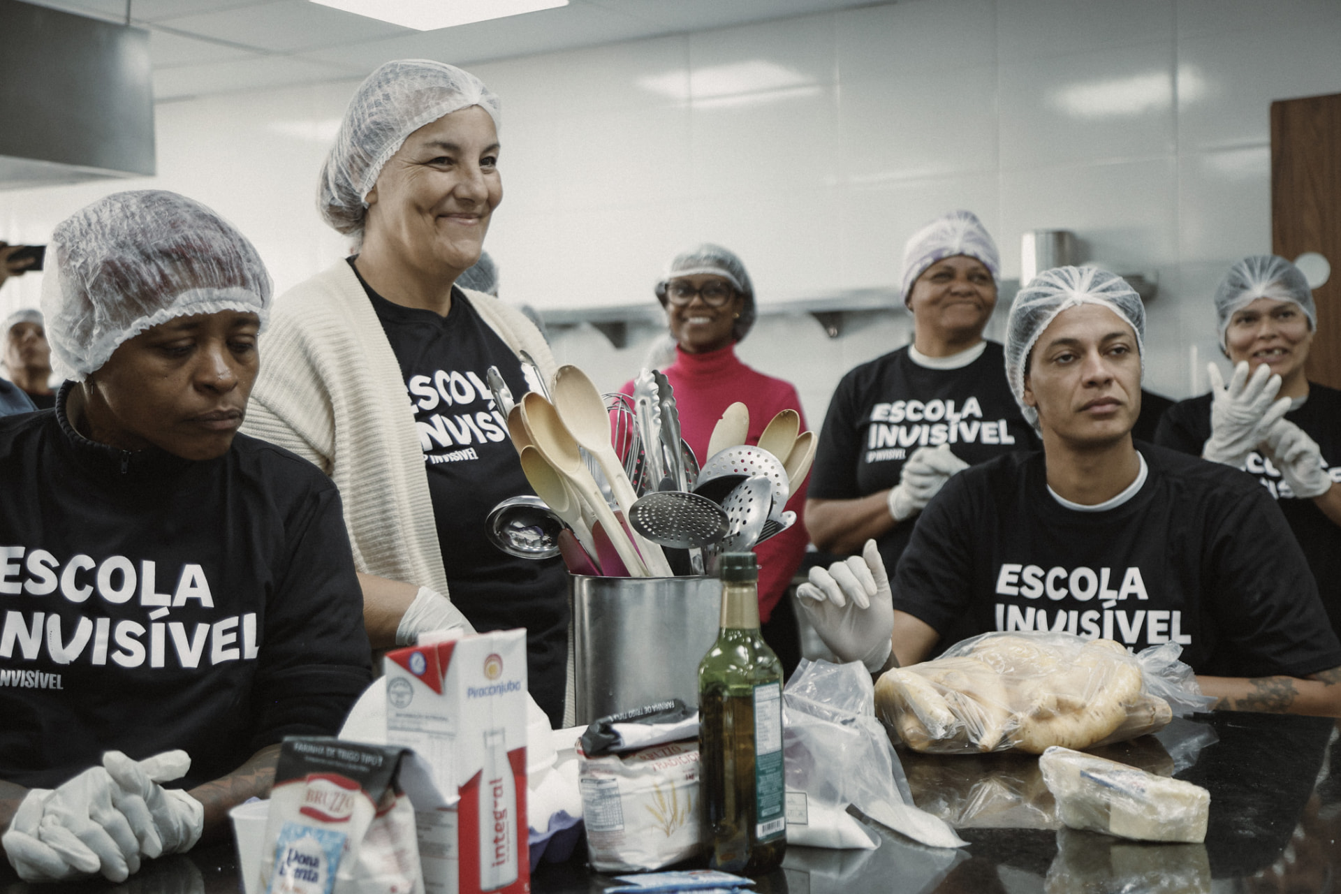 Alunas da Escola Invisível, de touca e camiseta do projeto, sorriem em uma aula de cozinha.