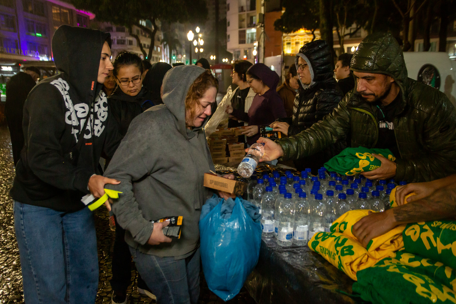 Voluntários distribuem marmitas, água e peças de roupa a pessoas em situação de rua no Largo do Paissandú, à noite.