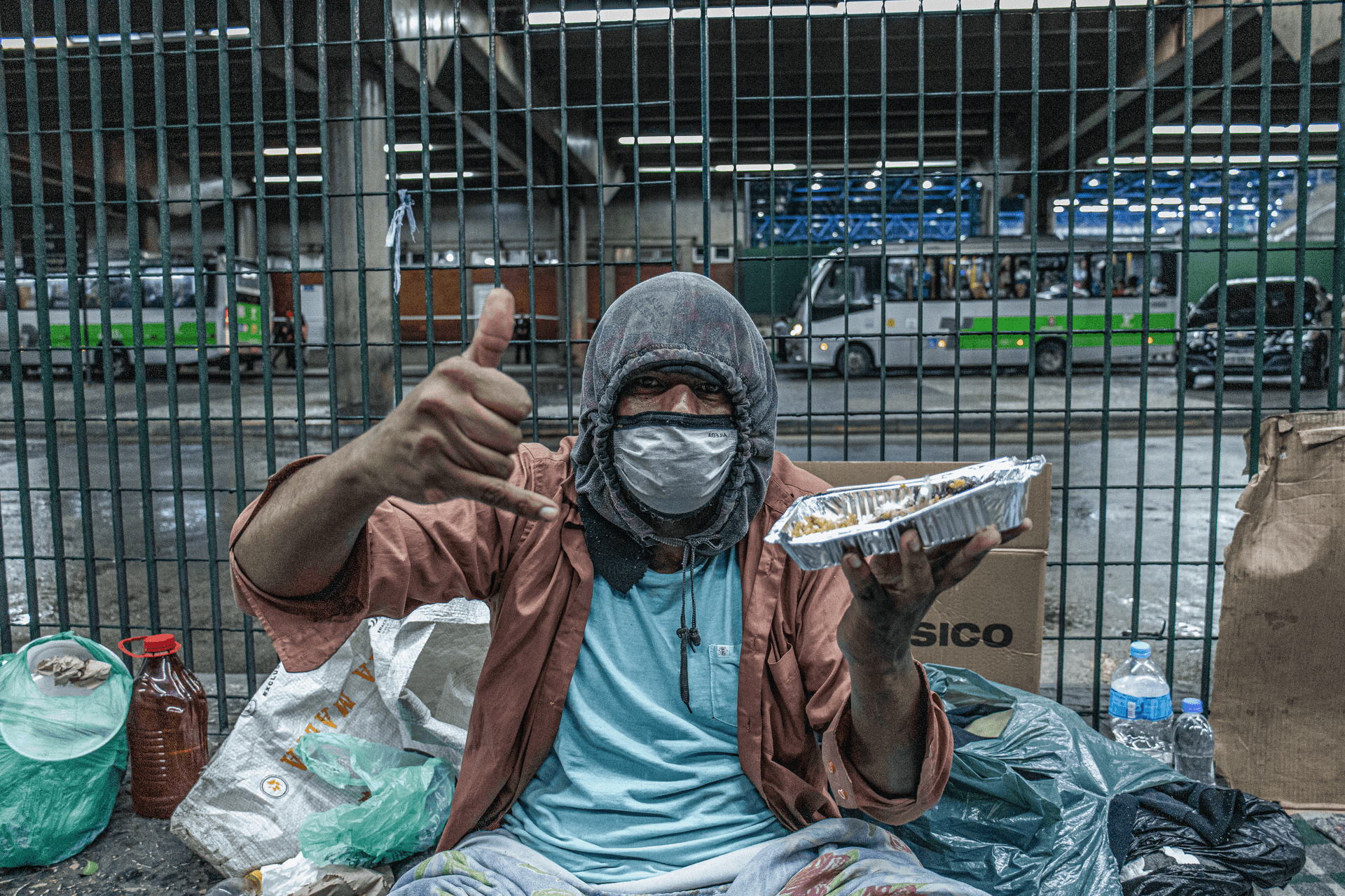 Foto de um homem em situação de rua segurando uma marmita. Ele está usando máscara facial, gorro de frio e faz sinal de 'hang loose' com a mão.