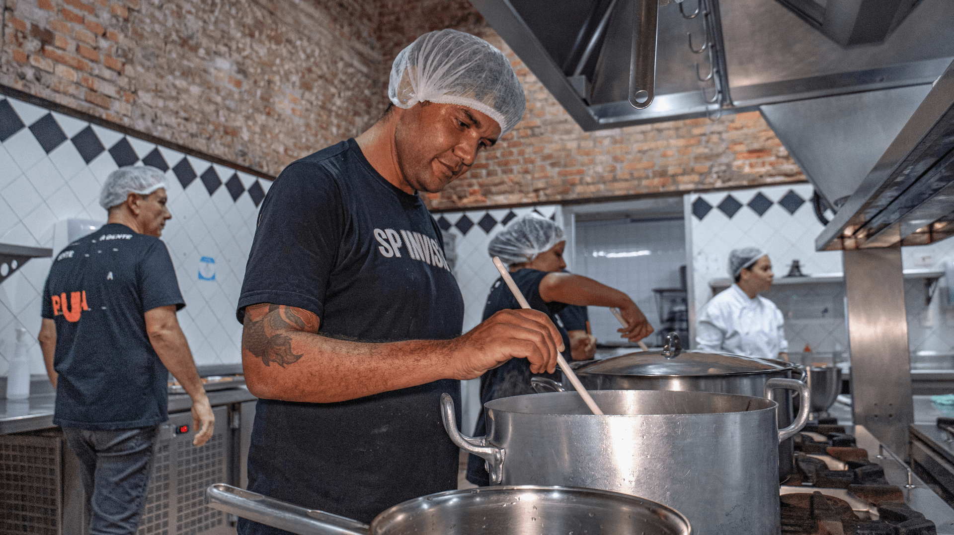 Alunos do curso Gastronomia Invisível em uma cozinha profissional. Um dos alunos usa touca descartável, camiseta da SP Invisível. Ele está mexendo em um caldeirão.
